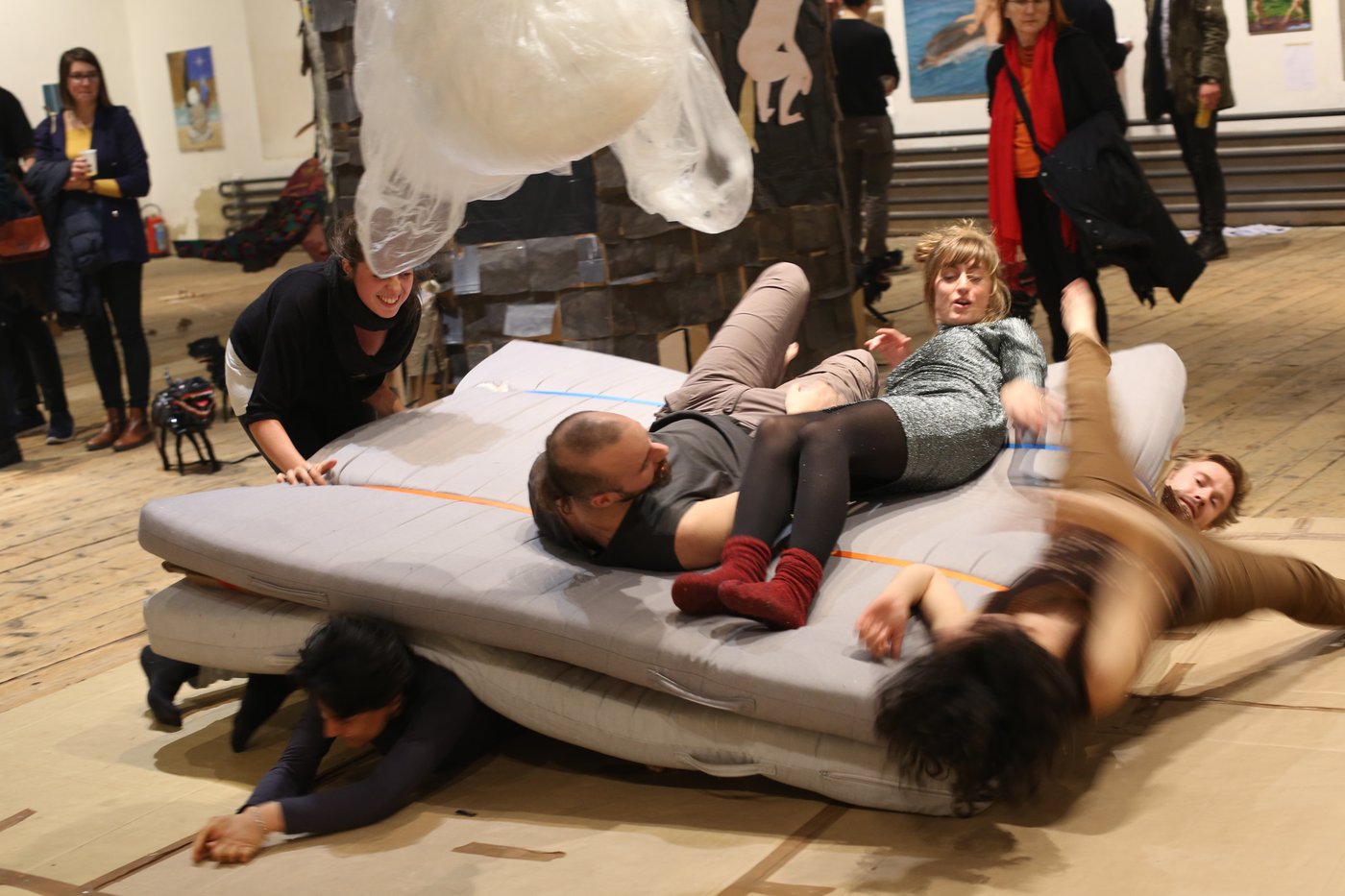 Young people are fooling around on a pile of mattresses. The situation seems very exuberant. In the background you can see some visitors of the tour and other works.