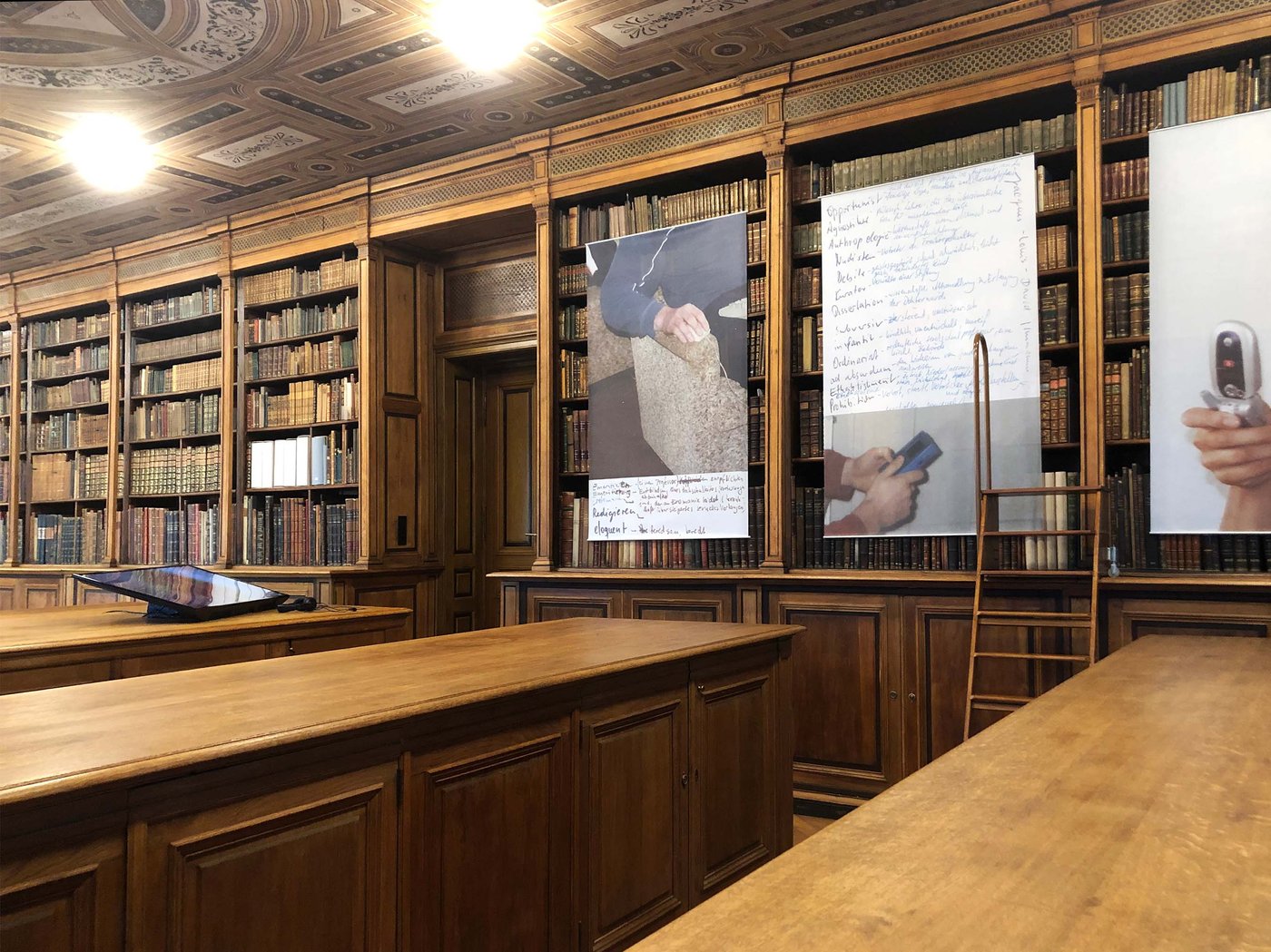 Exhibition view in the university library: on the wooden shelves hang exhibition objects in front of the books