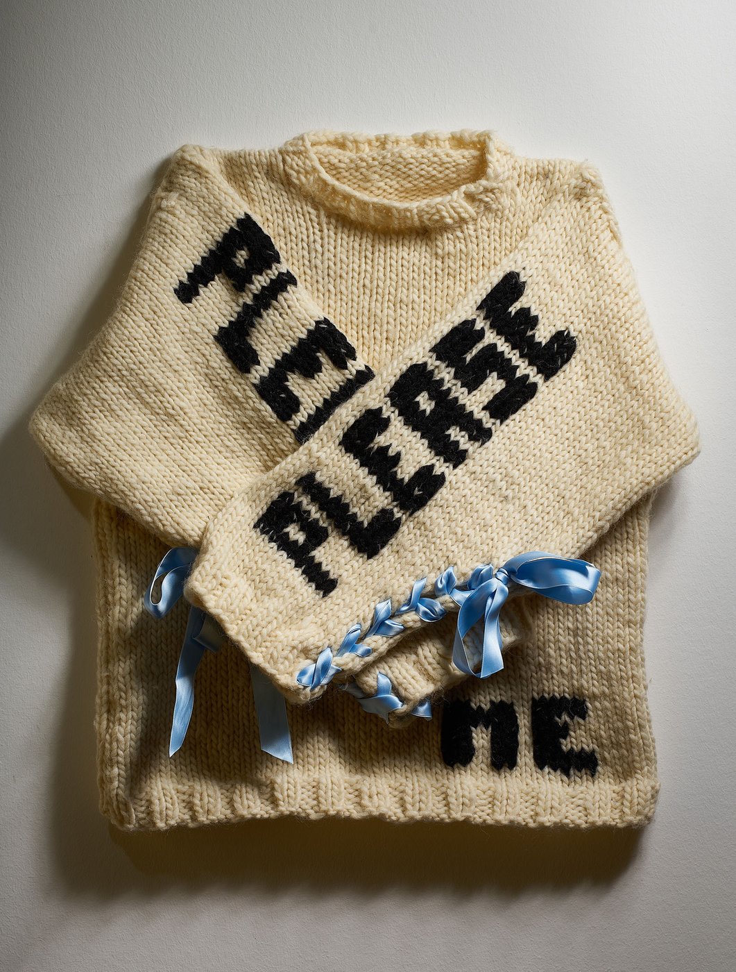 Seen is a cream-colored knit sweater with crossed arms. On each sleeve is embroidered the word "please", at the bottom – the word "me" (= please please me). On the sides the sleeves are decorated with blue ribbons.