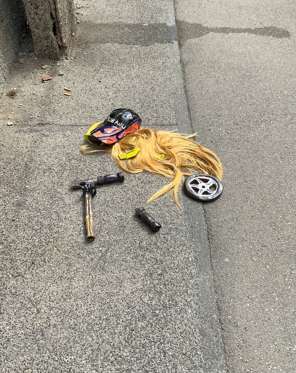 The picture shows a concreted sidewalk on which something happened. In the middle of the floor are fragments that tell a story. There is a wig with long glossy hair, on top of that a cap that reminds one of the the car-brand Subaru, the label of the brand is on it, decorated with tribal-style-flames. Underneath the cap and in the nest, that the hair has built on the street, are pieces of broken sunglasses. They have a neon color and one of the shards reveals the Gucci logo on them. Next to the human traces, are parts of a scooter. A handlebar from which one side has broken off, one other pole with a holder to regulate the size of the scooter and a reflecting metal wheel.The title of the artwork is: “Cute babe fell off the scooter and broke their Gucci glasses. Uff.“ All the pieces, accept for the wig, are made out of ceramics and have the same glossy, smooth surface the glaze creates.