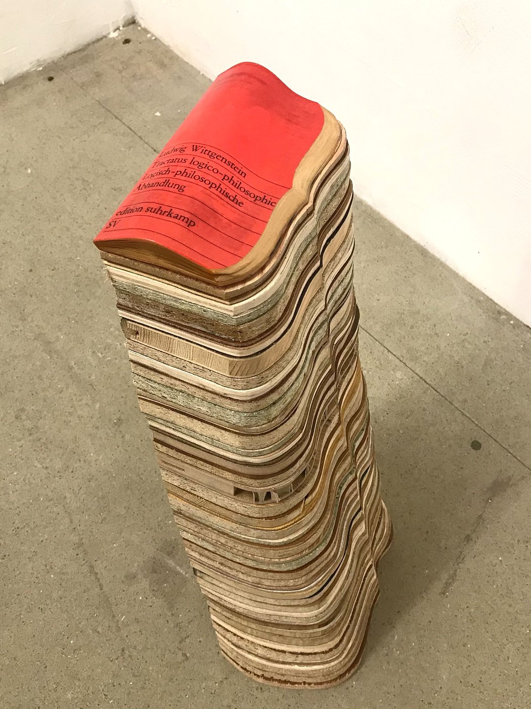 On display is a 65 cm high tower built from 50 layers of wood. The individual layers stand out visually from each other due to their different colours and types of wood. The top layer shows the cover of Wittgenstein's book “Tractatus logico-philosophicus” from edition suhrkamp in red. The tower was milled along the right side in the form of a curved bracket. The photo was taken from diagonally above right.