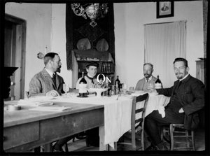 The black-and-white photograph shows four persons, three read as male and one as female, sitting at a long coarse wooden table eating and drinking wine. It is a scene from the 19th century. In the center of the picture the word ‘On’ can be read.