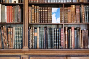 The color photograph in landscape format shows a section of a bookshelf with historical holdings, designed by Theophil von Hansen for the reading room of the Academy Library. Some books have been removed, and their place between the spines is taken by a portrait-format color photograph with a cloudy sky.