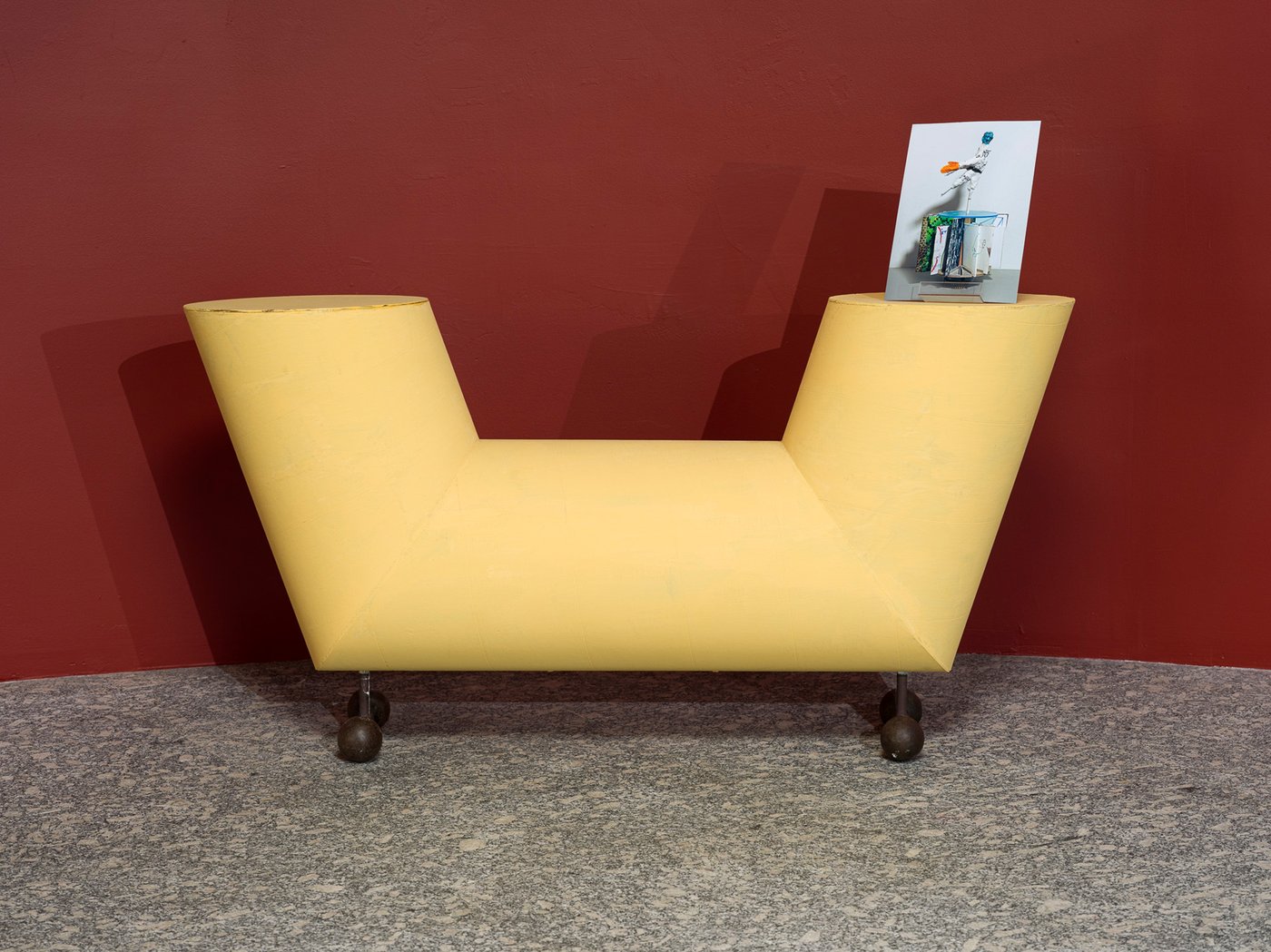 The color illustration in landscape orientation shows a yellow sculptural object that vaguely resembles a bench. On the right “armrest” stands a photograph depicting another small sculpture.