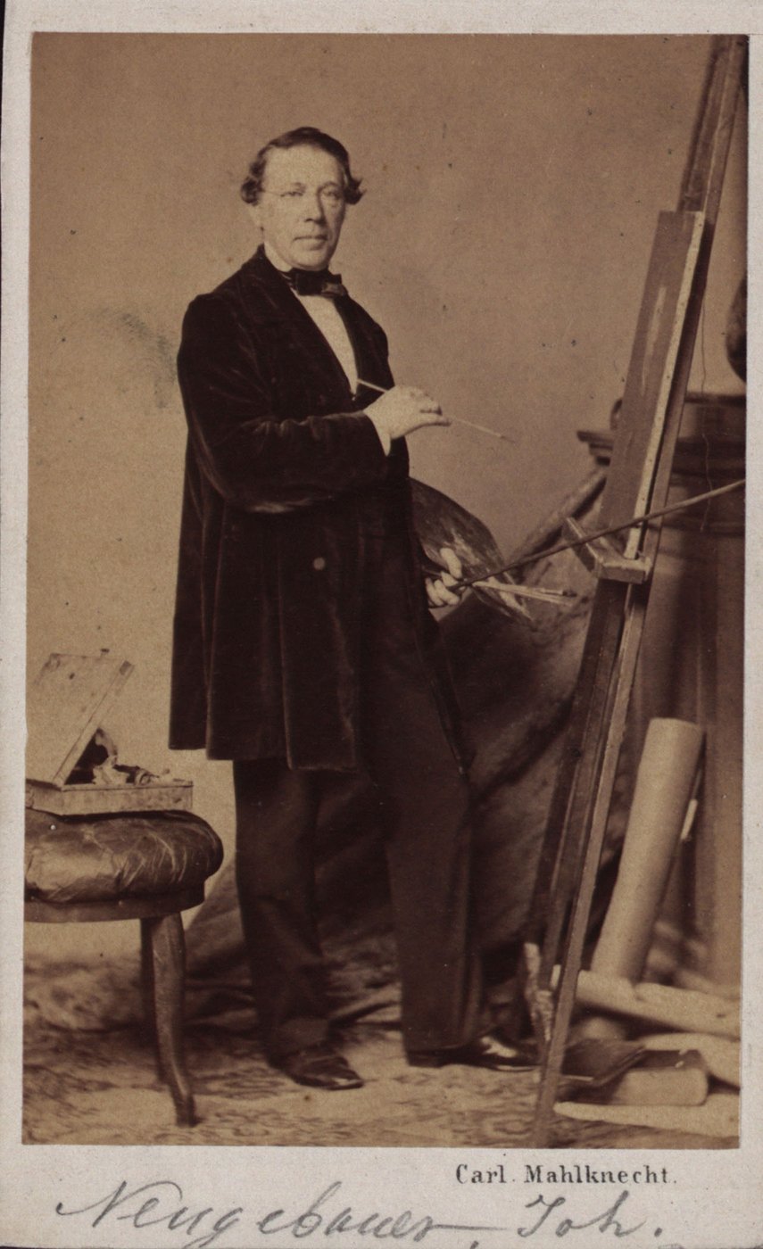 B&amp;W photograph by Joseph Neugebauer in portrait format. The painter in dark trousers and a long velvet jacket stands in front of an easel, holding a brush in his right, slightly raised hand and a palette and other brushes in his left. He looks into the camera.