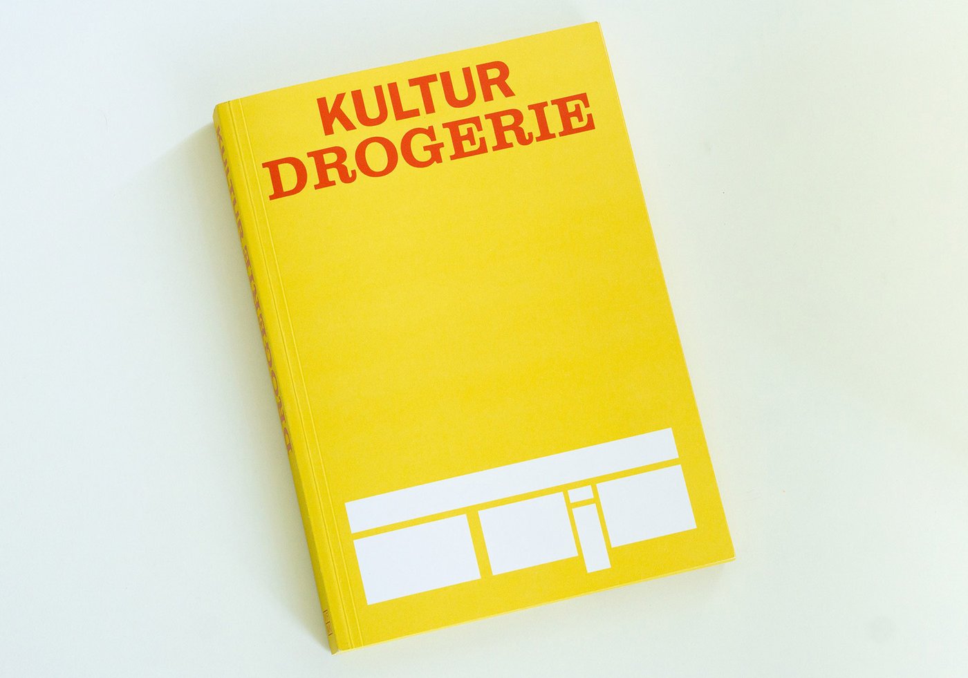 Color photo in landscape format: The “Kulturdrogerie” catalog, published in 2025 to mark the 20th anniversary of the artist-run space, lies on a light-colored surface. The yellow cover features the words “Kultur” and “Drogerie” in red capital letters, centered at the top and arranged one below the other. At the bottom is a white graphic consisting of a wide horizontal bar, with three rectangles below it; between the second and third rectangles is an upside-down exclamation mark.