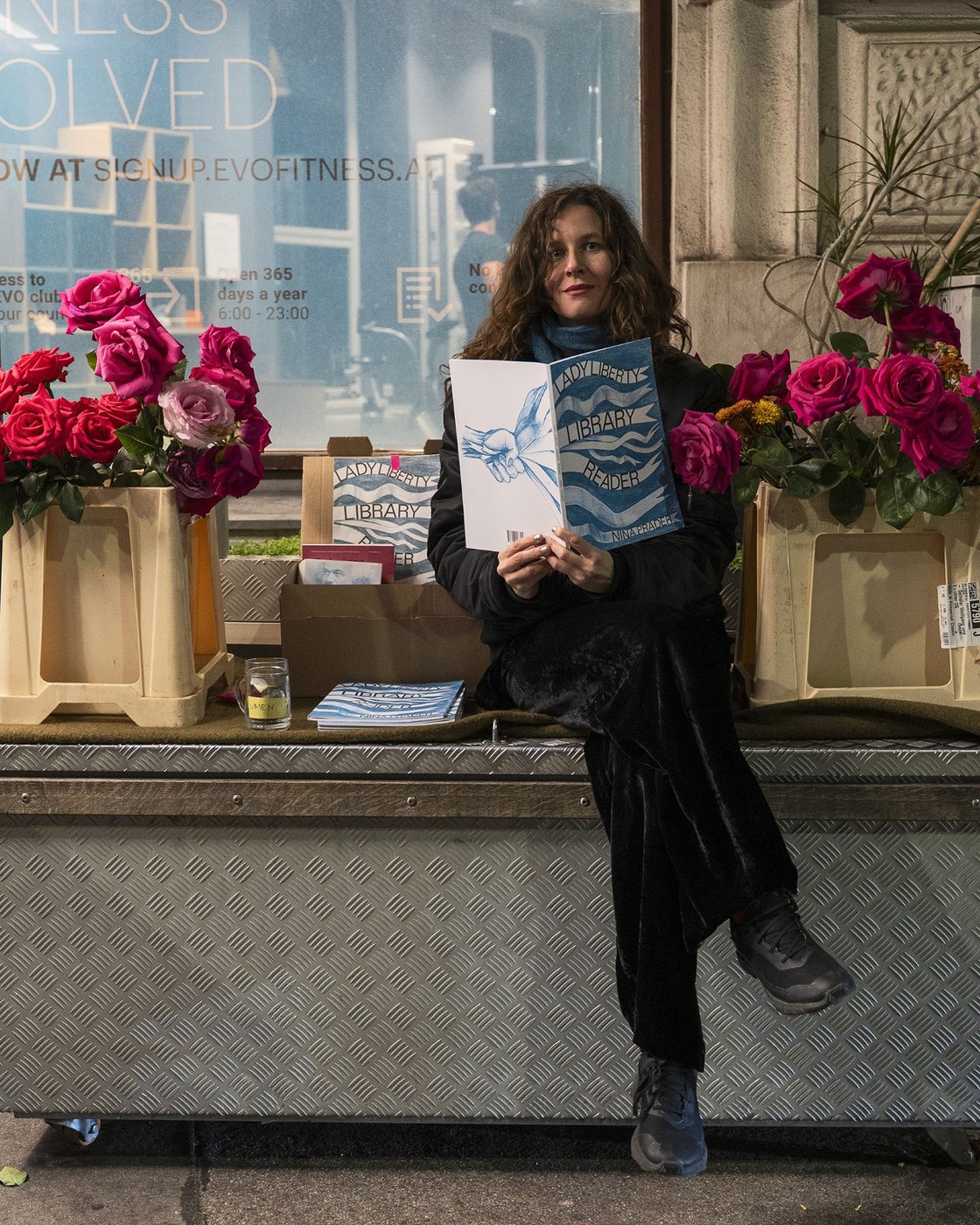 The color photograph, in portrait format, shows Nina Prader with her “Lady Liberty Library Reader.” She is sitting on a large metal rolling cart, her legs crossed, between a box of books and two plastic containers filled with roses in full bloom.